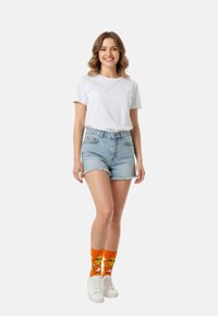 Witte crew-neck T-shirt, high-waisted denim shorts, oranje sokken met cartoongezichten, en witte sneakers op een neutrale achtergrond.
