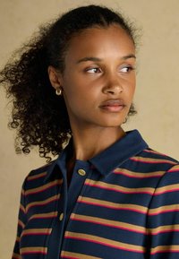 Polo tričko s námorníckymi pruhmi v ružovej a béžovej farbe, s golierom a zlatými akcentovými gombíkmi. Model má kučeravé vlasy.