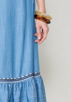 Vestido de chambray azul claro con dobladillo con volantes y acento de bordado floral. Lleva múltiples pulseras translúcidas en la muñeca.