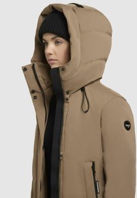 Giacca puffer khaki con un ampio cappuccio, dettagli neri, chiusura con zip e tasche frontali. Presenta una patch circolare con logo sulla manica.