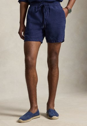 Mann trägt marineblaue Shorts mit Kordelzug und passende marineblaue Espadrilles, steht mit einer Hand in der Tasche.