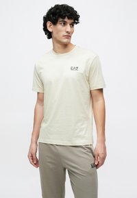 Gaiši bēša kokvilnas T-krekls ar īsām piedurknēm un apaļu kakla izgriezumu, ar nelielu melnu EA7 logo kreisajā krūtīs.