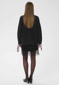 Un pull en tricot noir à coupe ample avec des liens sur les côtés, superposé à une jupe noire ornée de sequins, longueur genoux, associée à des talons noirs.