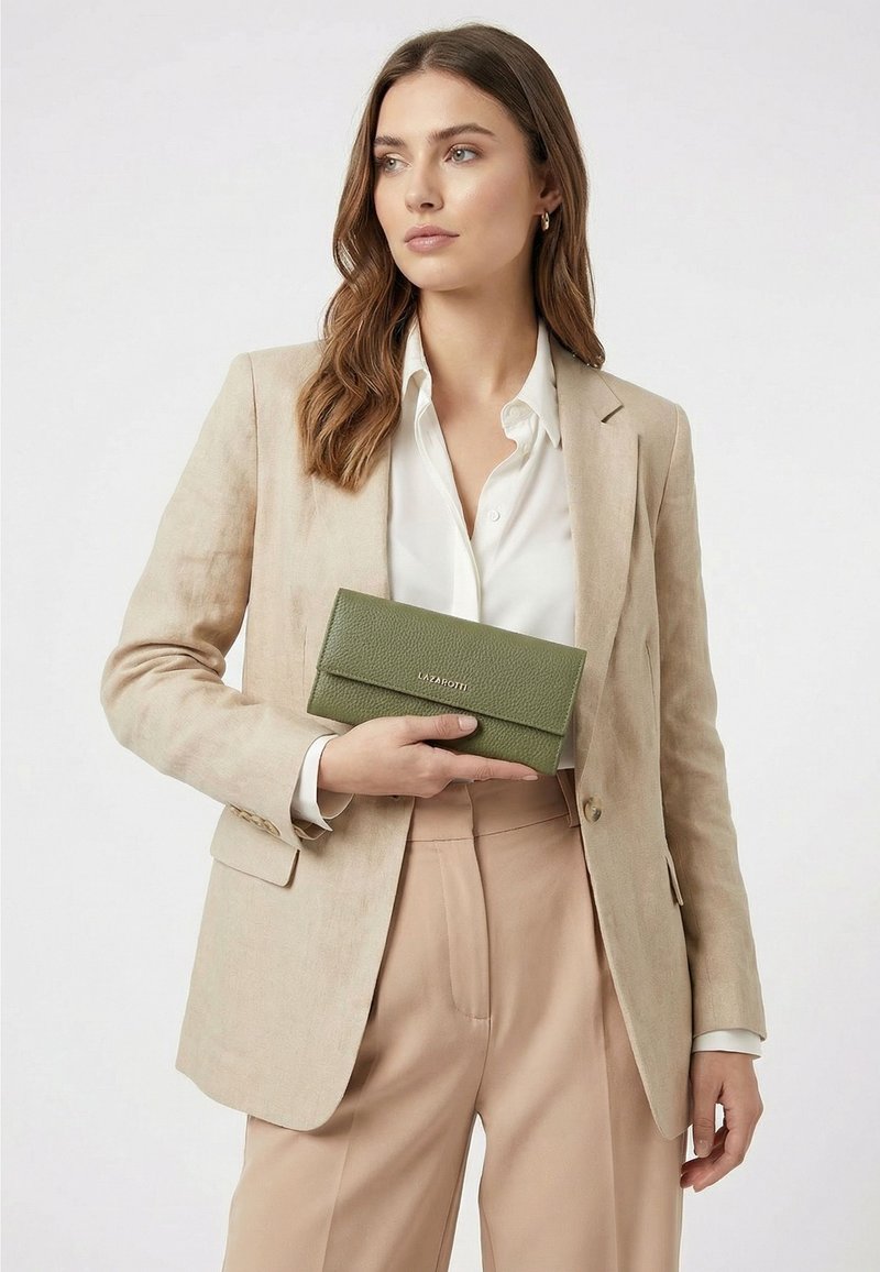 Mujer con blazer y pantalones beige sostiene una cartera de cuero verde con el logo Lazarotti sobre fondo blanco.