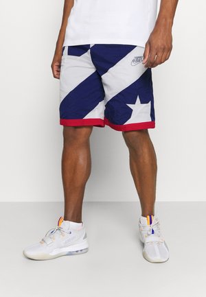 Mann trägt Nike-Shorts in Blau, Weiß und Rot mit Stern-Design, weiße Sportschuhe mit orangefarbenen und violetten Akzenten, steht vor weißem Hintergrund.