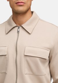 Beige lynlåsjakke med krave, tekstureret stof, to brystlommer og en glat lynlås. Stoffet har et diskret diagonalt mønster.