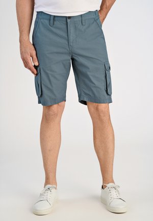 Man draagt blauwe cargoshorts met zijzakken en witte sneakers, staand tegen een effen lichte achtergrond.