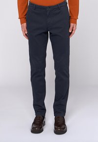 Man som bär smala mörkblå byxor med bruna loafers och en orange långärmad skjorta, stående framför en vit bakgrund.