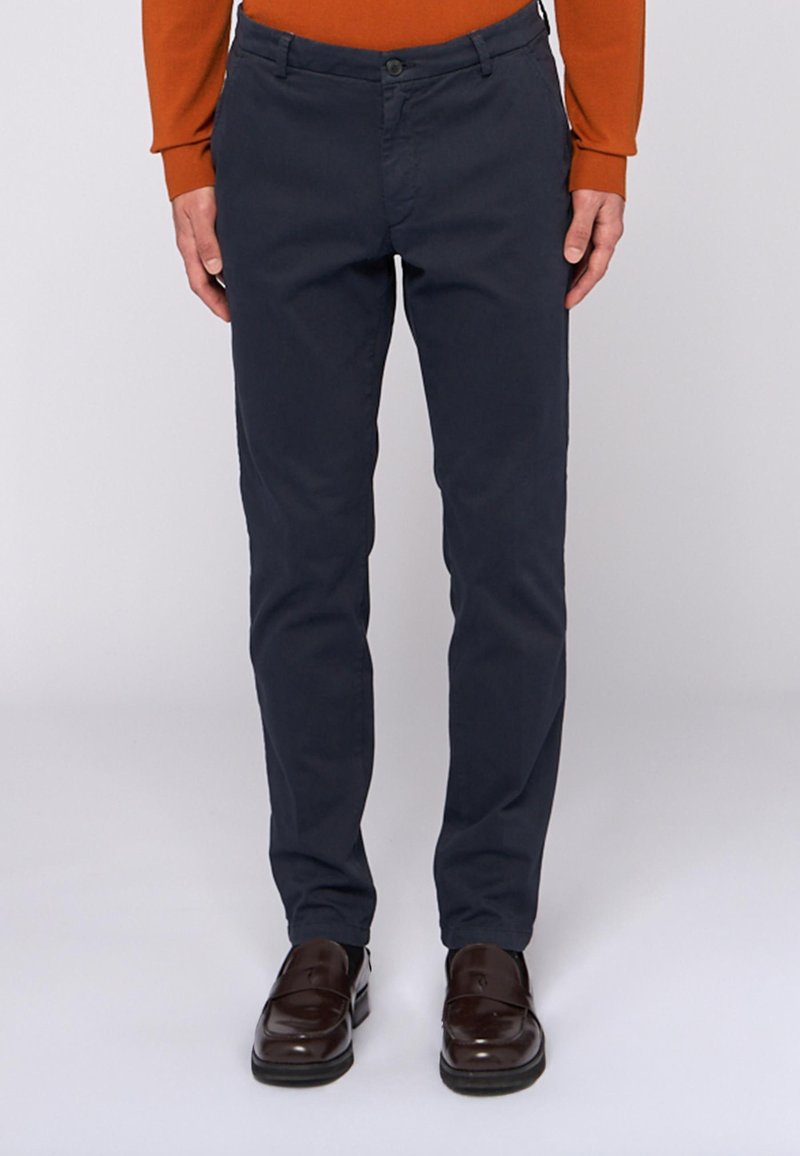 Man som bär smala mörkblå byxor med bruna loafers och en orange långärmad skjorta, stående framför en vit bakgrund.