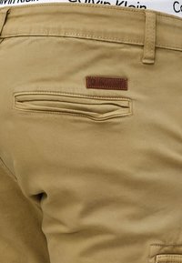 Beige chinos with a smooth texture, featuring a back pocket and a brown leather brand label. Stitched detailing on waistband and pockets.