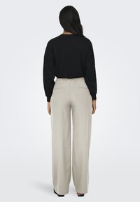 Donna con lunghi capelli neri che indossa una maglietta nera a maniche lunghe e pantaloni beige a gamba larga, in piedi con la schiena rivolta verso la fotocamera.