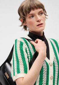 Πλεκτή ζακέτα με ρίγες σε πράσινο και κρεμ χρώμα, με βαθύ V λαιμόκοψη και διακοσμητικά κουμπιά, σε συνδυασμό με μια μαύρη, λείου υφής τσάντα.