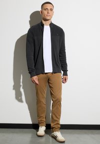 Man som bär en svart zip-up tröja, vit T-shirt, bruna byxor och vita sneakers, står mot en vit vägg och kastar en skugga.