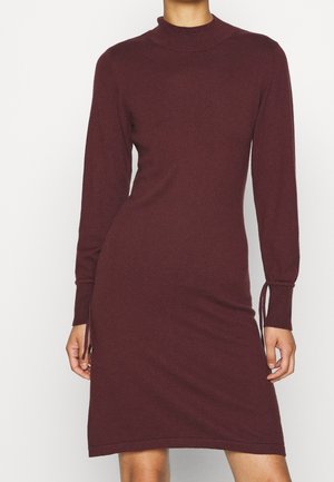 Getailleerde wijnrode jurk met lange mouwen tot op de knie, met col en strikdetails aan de mouwen, gedragen door een staand persoon tegen een effen achtergrond.