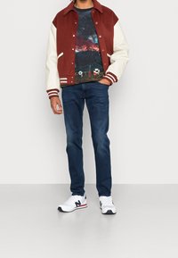 Rode en witte varsity-jack, grafisch T-shirt met een nachtlandschap, blauwe jeans en witte sneakers met zwarte accenten.