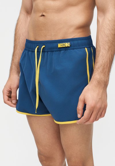 Άνδρας χωρίς πουκάμισο που φοράει μπλε μαγιό με κίτρινη κορδονάκι, περιθώριο, πλευρικές ρίγες και λογότυπο "Moschino" στη ζώνη μέσης.