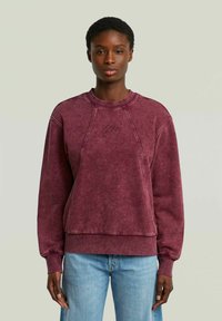 Felpa bordeaux a manica lunga con maniche raglan, texture sbiadita e logo ricamato sul petto, abbinata a jeans azzurri chiari.
