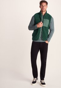 Man wearing green fleece vest over gray long-sleeve shirt, black pants, and black sneakers with white soles and laces, standing with one hand in pocket.