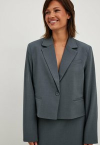 Blazer grigio su misura con chiusura a un bottone, rever a punta e due tasche frontali; realizzato in tessuto liscio e strutturato.