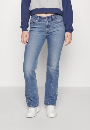 Osoba ubrana w niebieskie spodnie typu straight-leg z plamami, białe trampki oraz szaro-granatową krótka bluzę z długim rękawem, stojąca na tle białego tła.