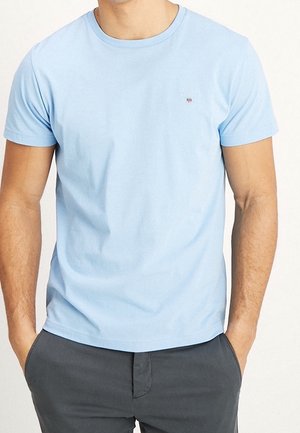 Man wearing a light blue short-sleeve t-shirt with dark gray pants, hands casually in pockets, plain white background.