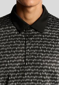 Svart och gråmönstrad pikétröja med en mjuk krage, med knappslå och en slät textur. Anmärkningsvärd design inkluderar ett geometriskt mönster.