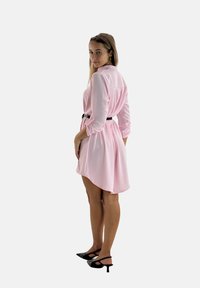 Roze, knopenjurk met lange mouwen, vastgebonden in de taille met een zwarte riem. Glad materiaal en een geflarde, licht asymmetrische zoom.