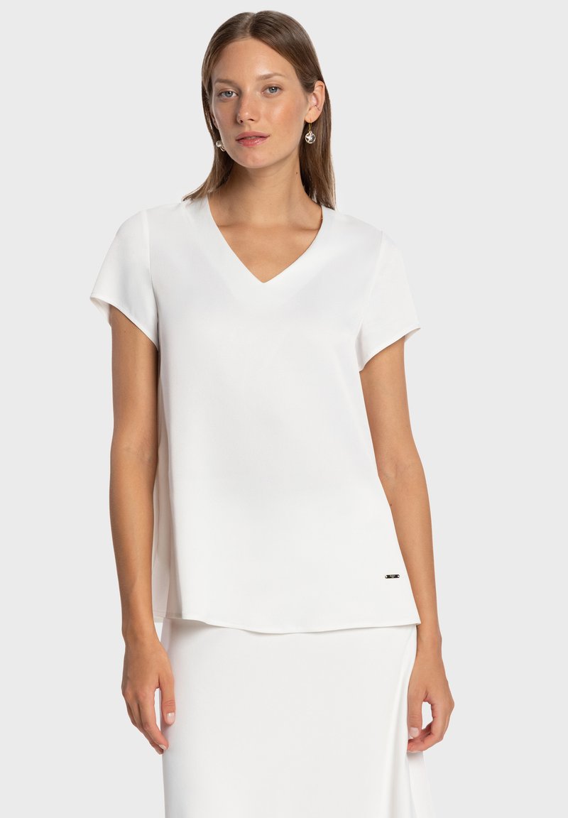 Blouse blanche à manches courtes avec col en V, texture lisse, coupe décontractée et léger drapé. Comprend un petit accent de marque à l'ourlet.