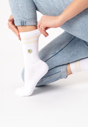 Person trägt hellblaue Jeans und weiße, gerippte Socken mit gelben Streifen und einer kleinen bestickten Zwiebel und richtet die Socke.
