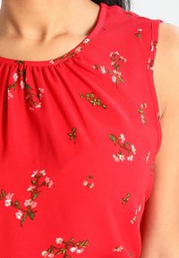 Top senza maniche rosso con scollatura arricciata, decorato con ricami floreali bianchi e verdi su un tessuto liscio.