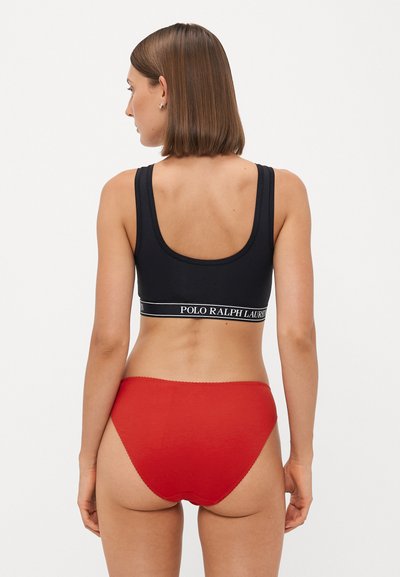 Black sports bra with a wide branded band, paired with high-waisted red briefs. The fabrics are smooth with minimal stitching details.