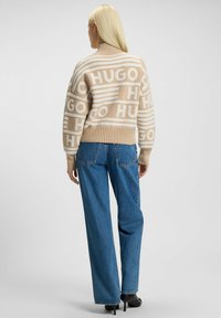 Beige trui met herhaaldelijk "HUGO" patroon, hoge kraag en geribde zoom. Gecombineerd met wijde blauwe jeans en zwarte hakken.