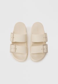 Sandali slide beige con due fasce larghe e fibbie rettangolari. Texture liscia e suole piatte. Design minimalista.