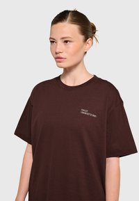 Ung kvinna med brunt hår uppsatt i en tofs, klädd i en lös mörkbrun T-shirt med liten vit text på vänster övre sida.