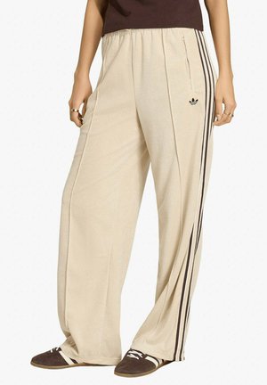 Persona che indossa pantaloni sportivi Adidas beige con strisce nere e logo, abbinati a sneakers marrone scuro con lacci bianchi e strisce.
