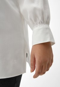 Witte button-up blouse met elastische manchetten, zijslitsen en een klein geborduurd logo aan de zoom. Glad materiaal en een getailleerd ontwerp.