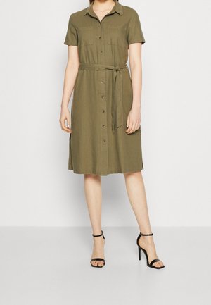 Robe-chemise à manches courtes de couleur vert olive avec fermeture à boutons sur le devant, ceinture à nouer à la taille et deux poches poitrine. Associée à des talons noirs à brides.