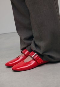 Rode patenten leren schoenen met een spitse neus en zijopeningen, vastgemaakt met een brede band met een zilveren gesp, gecombineerd met grijze broek.