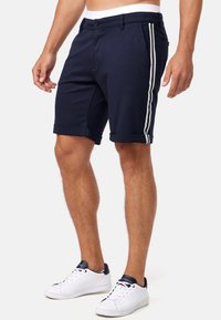 Mørkeblå shorts laget av et mykt stoff, med en skreddersydd passform, rullekanter og to hvite sidelister for kontrast.