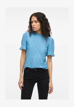 Nainen, jolla on kiharat hiukset, pukee ylleen vaaleansinisen pitsipaidan puffihauerilla ja mustat korkeavyötäröiset farkut, katsoen sivulle.