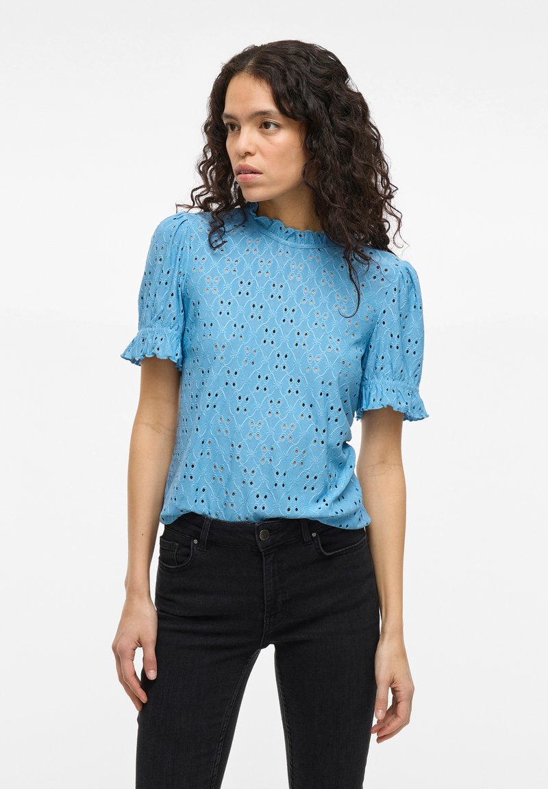 Lyseblå bluse med brodert hullmønster, korte volangermer og scallopkrage, kombinert med svarte jeans.