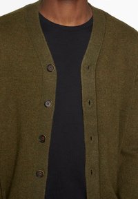 Olivgrön stickad cardigan med V-ringning, prydd med fem bruna knappar och ribbade kanter, lager ovanpå en svart skjorta.