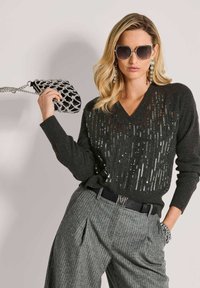 Frau trägt einen schwarzen Paillettenpullover, graue Nadelstreifenhose, große Sonnenbrille, hängende Ohrringe und hält eine schwarze und silberne Clutch.