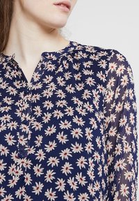 Marinblus med vitt prästkrage-mönster och röda mittar, med en knappslå och mjukt, genomskinligt tyg. Subtila veck nära halsringningen.