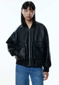 Schwarze Leder Bomberjacke mit Reißverschluss, zwei Fronttaschen und gerippten Bündchen. Übergrößenpassform und glatte Textur. Getragen mit blauen Jeans.