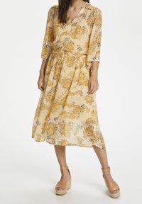 Abito floreale giallo con maniche a tre quarti, scollatura a V, vita arricciata e gonna fluida. Indossato con scarpe con plateau color beige.