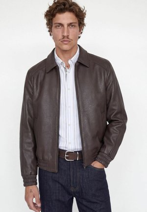 Homme portant une veste en cuir marron foncé sur une chemise blanche rayée et un jean foncé, debout avec une main dans la poche sur fond blanc.