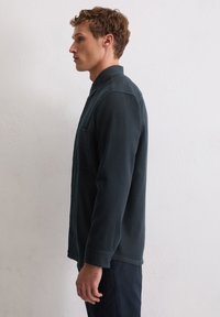 Dunkelgrünes Baumwollhemd mit Kragen, langen Ärmeln, Brusttasche und knöpfbaren Manschetten, seitlich an einem Modell gezeigt.