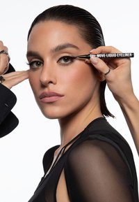 Nestemäinen eyeliner mustassa putkessa, jossa lukee "ONE MOVE", levitetään mallin silmälle. Mallilla on sileä ihonväri ja hänellä on päällään musta, läpinäkyvä toppi.