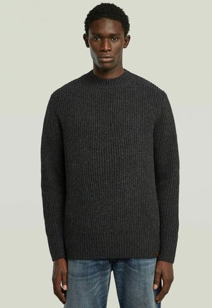Junger Mann, der einen dunkelgrauen, gerippten Strickpullover und blaue Jeans trägt und vor einem schlichten, hellen Hintergrund steht.